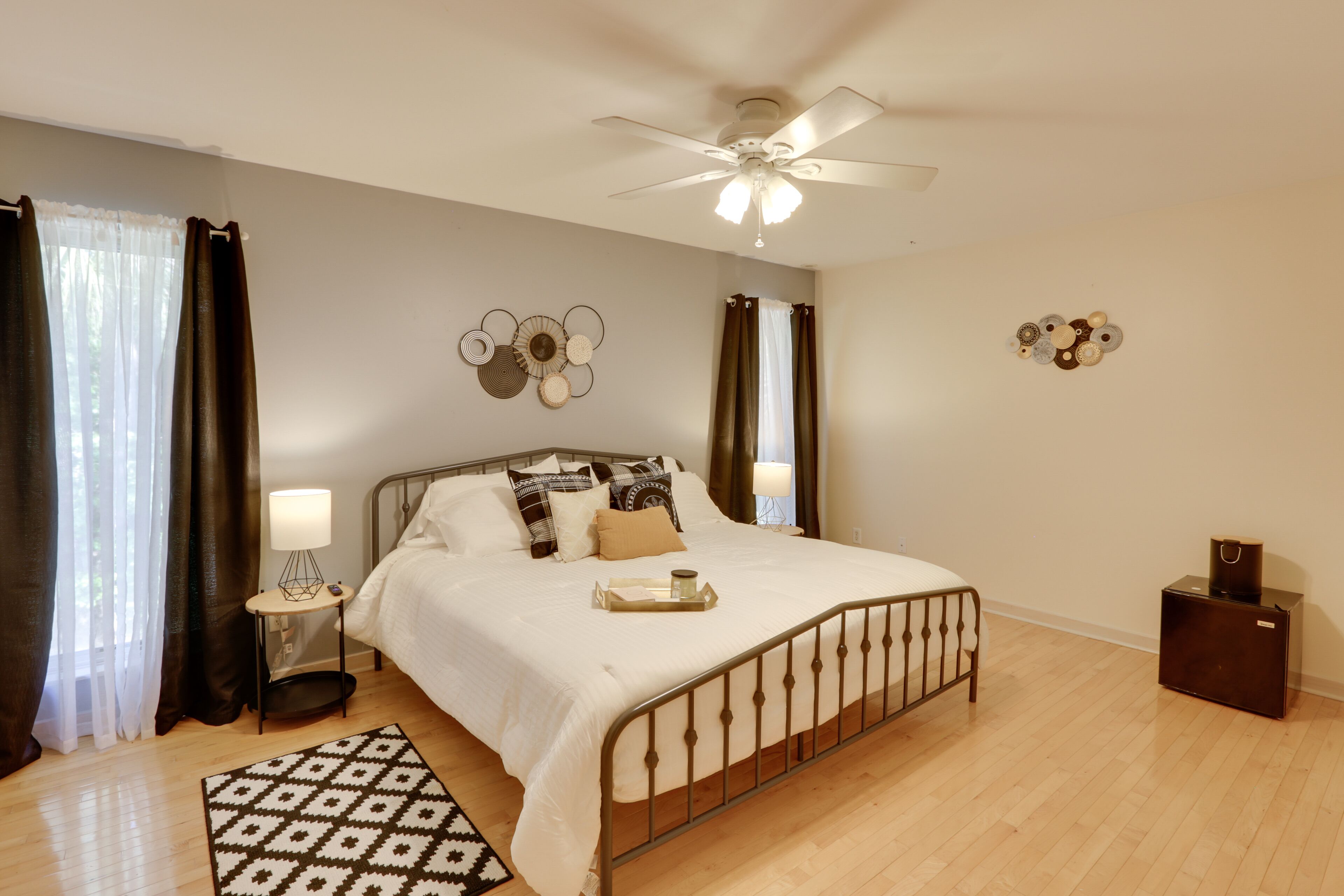 Een slaapkamer met een bed, een plafondventilator, een nachtkastje met een lamp, een bijpassend vloerkleed en decoratieve wandkunst.