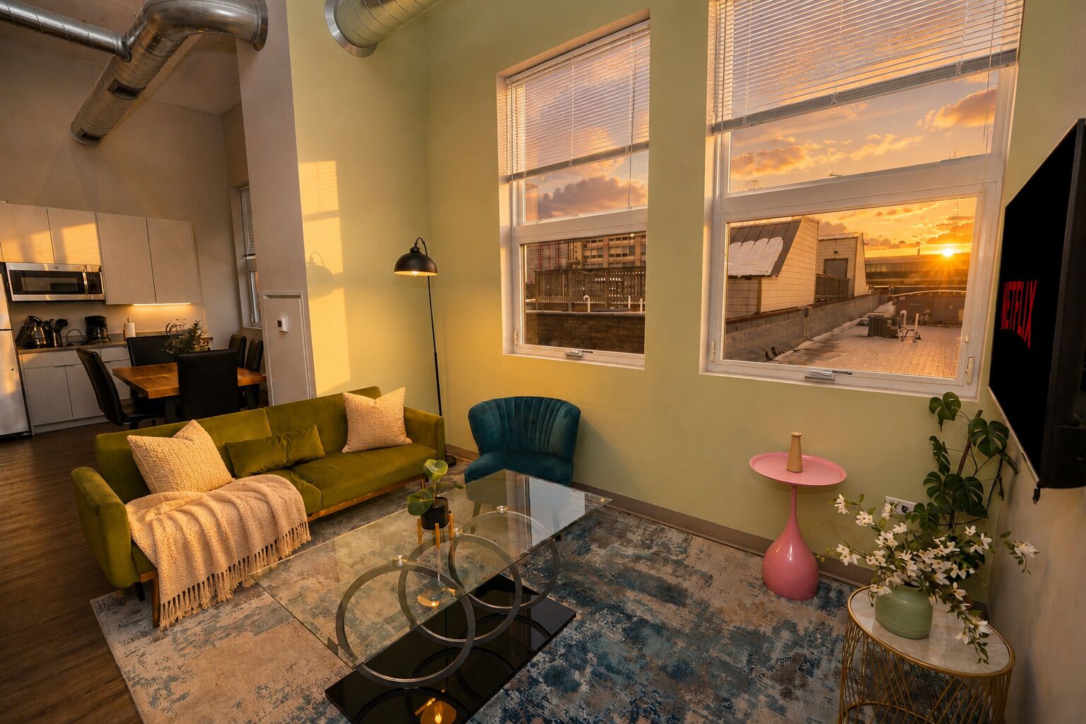 A modern living room with a green sofa, a blue armchair, and a coffee table. There are two windows with blinds, a pink side table, and a plant.
