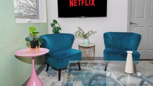 A living room with teal armchairs, a pink table, a glass coffee table, and a TV displaying "NETFLIX".