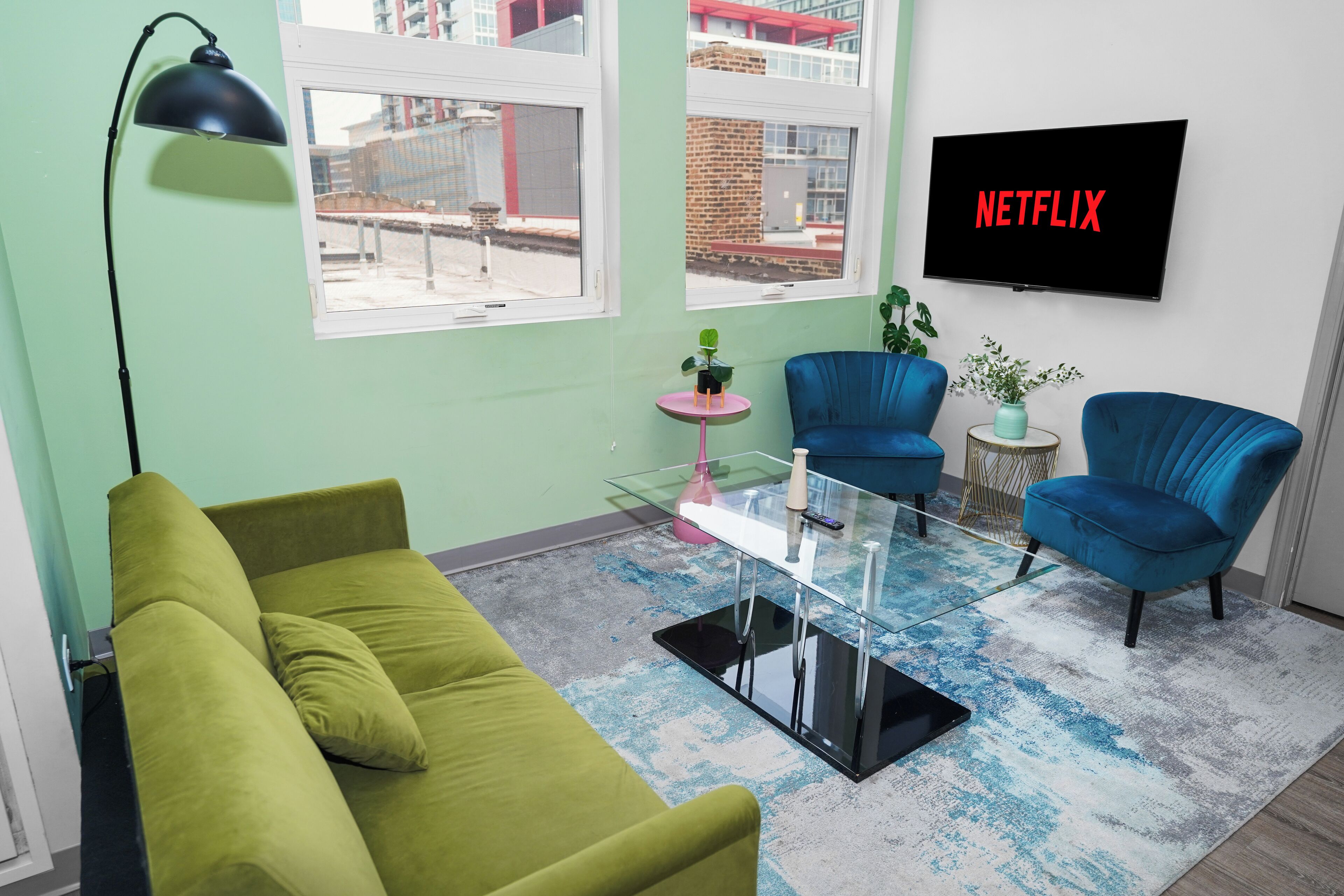A living room with a green sofa, two blue armchairs, a glass coffee table, a floor lamp, a TV displaying 'NETFLIX', and a view of a construction site through the window.