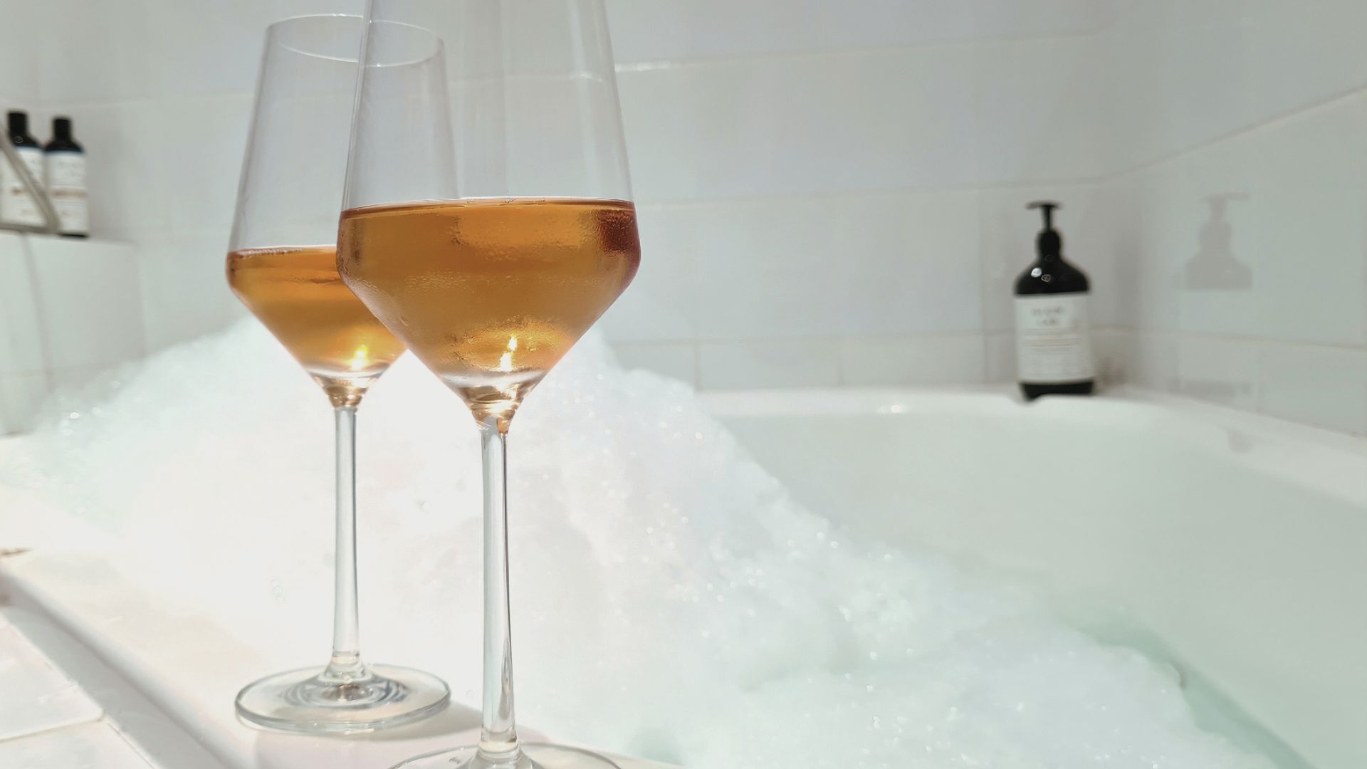 Two glasses of champagne in a bathtub filled with bubbles.