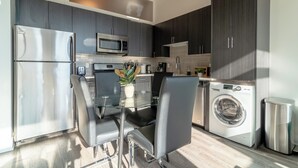 Une cuisine moderne dotée d’appareils électroménagers en acier inoxydable, d’une table à manger en verre et de meubles de rangement aux couleurs sombres.