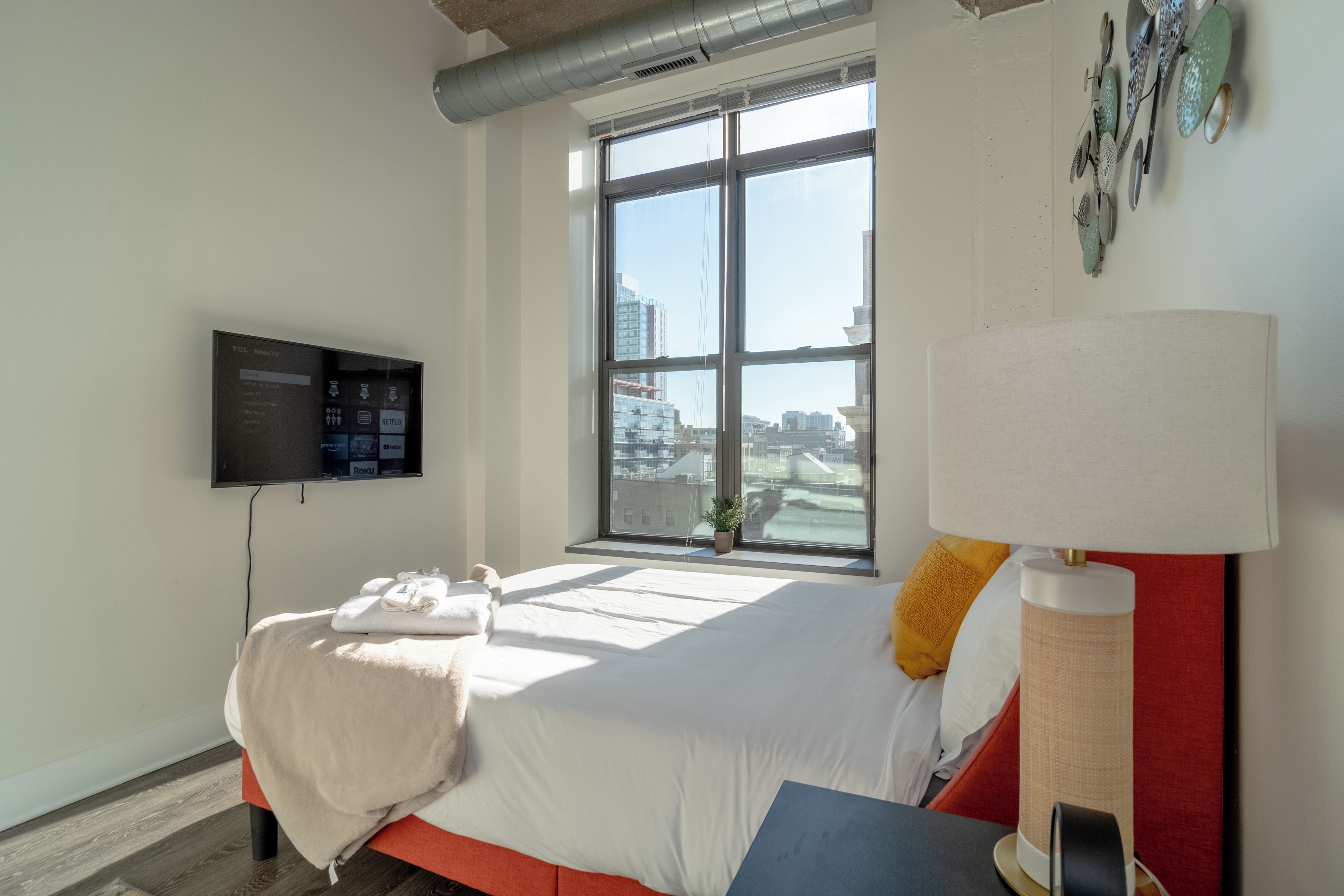 A hotel room with a bed, a TV mounted on the wall, a window with a view of the city, and a lamp on the bedside table.