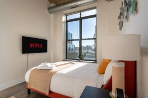 A hotel room with a bed, a TV displaying 'NETFLIX,' a window with a city view, and a lamp.