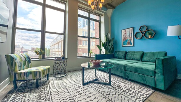 A living room with a green sofa, a patterned rug, and large windows offering a city view.