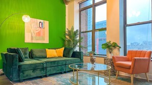 Un sofá verde con cojines amarillos, una mesa de centro redonda de vidrio y un sillón rojo en una habitación con ventanas amplias y vistas a la ciudad.