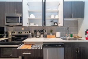 A modern kitchen with a built-in microwave, stove, and dishwasher. There is a wooden cutting board drawer and a variety of kitchen utensils.