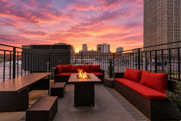 A rooftop terrace with a fire feature, seating area, and a view of the cityscape at sunset.