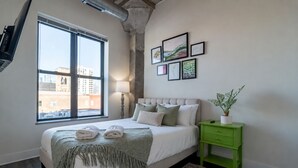 A bedroom with a bed, a window, a green nightstand, and a wall with framed artwork. - Premium 2BR Michigan Suite with Skyline Views Near McCormick Place & Lakefront (Chicago)