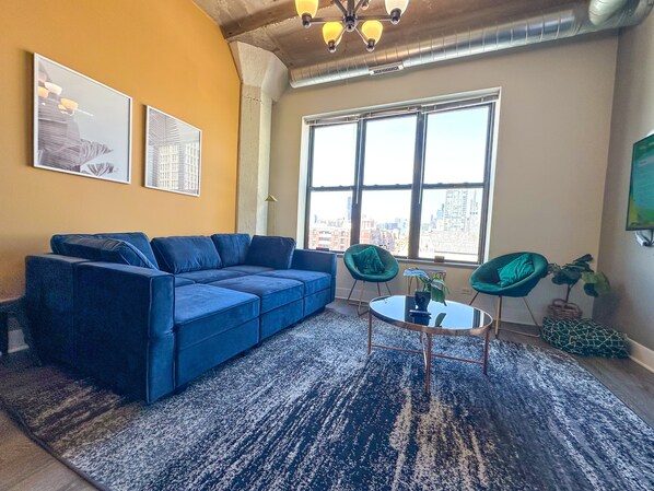 Ein Wohnzimmer mit einem blauen Sofa, zwei grünen Sesseln, einem kleinen Tisch und einem großen Fenster mit Blick auf die Stadt.