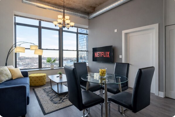 A modern living room with a glass table, black chairs, a sofa, a yellow ottoman, a TV on the wall, and a city view through large windows. - Bright & Stylish 2BR Family Unit Near McCormick Place & Grant Park (Chicago)