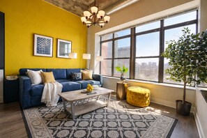 A living room with a blue sofa, a coffee table with fruit, a yellow ottoman, and a large window with a city view. - Bright & Stylish 2BR Family Unit Near McCormick Place & Grant Park (Chicago)