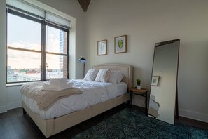 A bedroom with a large bed, a window with blinds, a bedside table with a lamp, and a mirror. - Bright & Stylish 2BR Family Unit Near McCormick Place & Grant Park (Chicago)