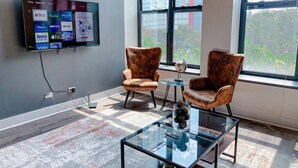 A modern office space with a large flat-screen TV displaying streaming services, two leather armchairs, a glass coffee table, and a view of a cityscape through large windows. - Modern 2BR Skyline View Near McCormick Place & Millennium Park (Chicago)