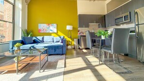 A modern living room with a blue sofa, a coffee table, and a dining area with a table and chairs. There is a kitchen area with cabinets and appliances. - Modern 2BR Skyline View Near McCormick Place & Millennium Park (Chicago)
