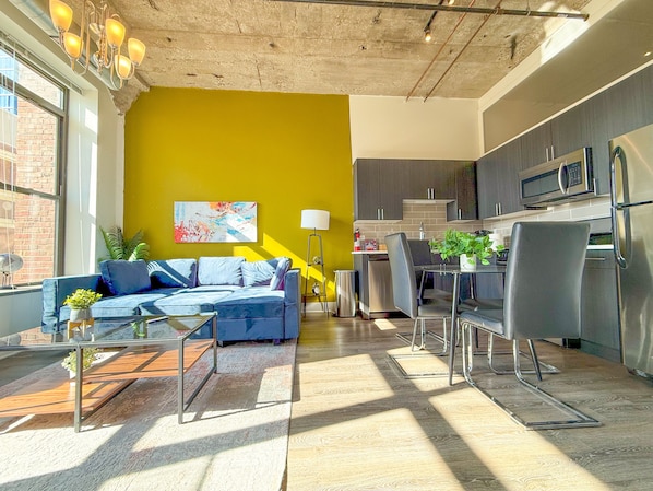 Un salon moderne avec un canapé bleu, une table basse et un coin repas comprenant une table et des chaises. Il y a également un coin cuisine avec des placards et des appareils électroménagers.