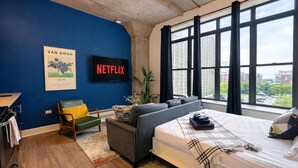 Ein modernes Schlafzimmer mit einem großen Bett, einem grauen Sofa, einem grünen Sessel und einem Fernseher, der 'NETFLIX' anzeigt.