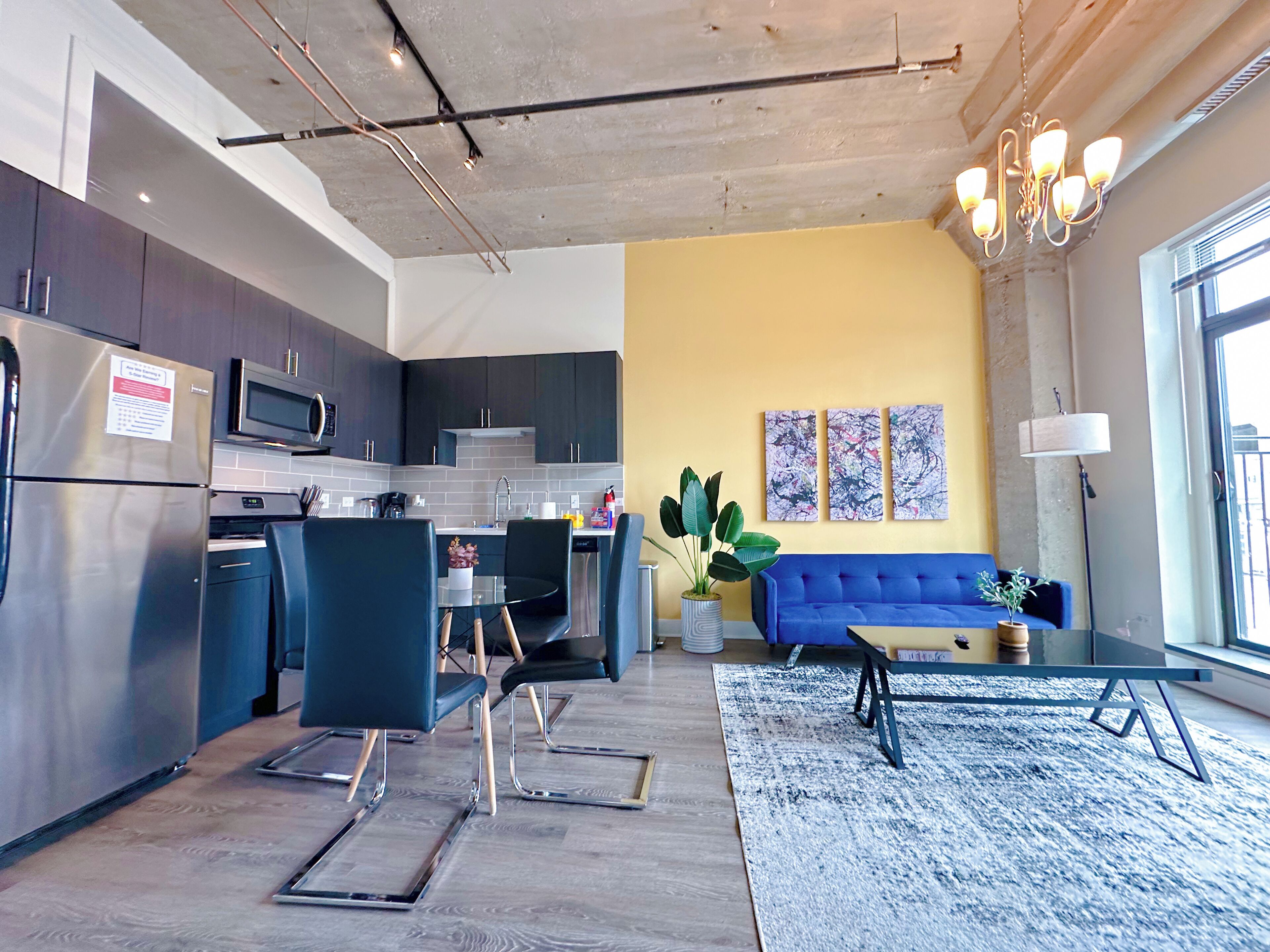A modern kitchen with dark cabinets, a stainless steel refrigerator, and a microwave. There is a dining area with blue chairs and a glass table. A blue sofa and a coffee table are in the living room, which has a yellow accent wall and abstract art.