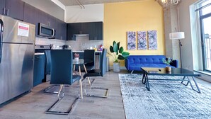 A modern kitchen with dark cabinets, a stainless steel refrigerator, and a microwave. There is a dining area with blue chairs and a glass table. A blue sofa and a coffee table are in the living room, which has a yellow accent wall and abstract art. - McCormick Place Patio skyline 2b/2b with optional Parking for up to 6 people (Chicago)