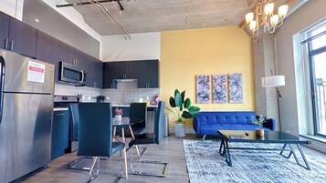 A modern kitchen with dark cabinets, a stainless steel refrigerator, and a microwave. There is a dining area with blue chairs and a glass table. A blue sofa and a coffee table are in the living room, which has a yellow accent wall and abstract art.