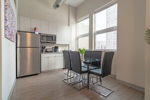 A modern kitchen with stainless steel appliances, a dining area with a glass table and chairs, and large windows offering a view of the city.
