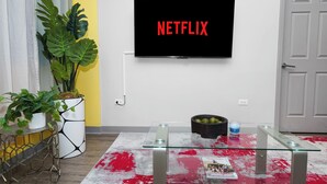 Un soggiorno con TV a parete che mostra il logo Netflix, un tavolino in vetro, una pianta in vaso e un tappeto con motivo rosso e bianco.