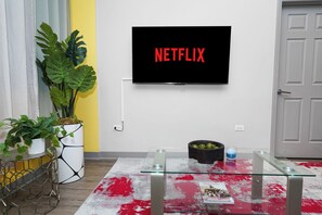 Sala de estar con un televisor montado que muestra el logotipo de Netflix, una mesa de centro de vidrio, una planta en maceta y una alfombra de diseño rojo y blanco.