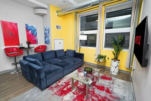 Una sala de estar moderna con un sofá azul, sillas rojas, una mesa de centro de vidrio y un amplio ventanal con cortinas.