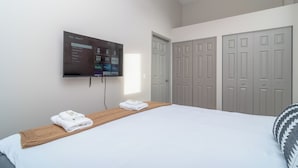 A hotel room with a bed, a mounted TV displaying a streaming service interface, a wooden bedside table with towels, and a set of grey doors. - Bright 2BR Apt with Gym and Optional Parking Near Soldier Field, McCormick Place (Chicago)