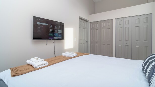 Camera d'albergo con un letto, una TV a parete che mostra l'interfaccia di un servizio di streaming, un comodino in legno con asciugamani e un insieme di porte grigie.