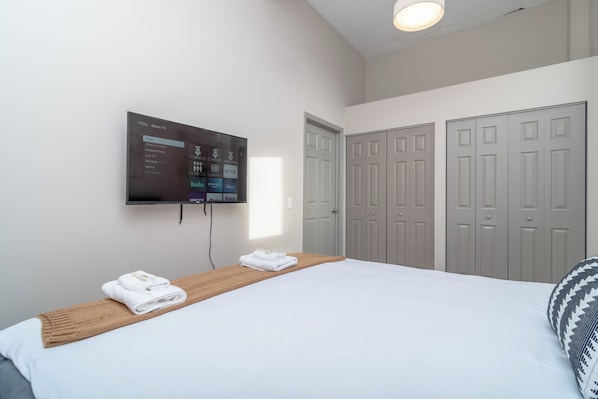 A hotel room with a bed, a mounted TV displaying a streaming service interface, a wooden bedside table with towels, and a set of grey doors.