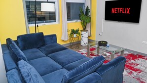 Una sala de estar con un sofá azul, una mesa de centro de vidrio, una planta en maceta y una pantalla de televisión mostrando "NETFLIX".
