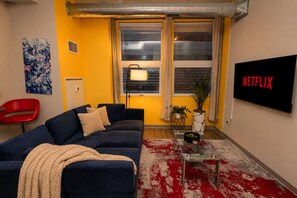 A living room with a blue sofa, a red chair, a TV screen with 'NETFLIX' on it, a window with curtains, and a red and white patterned rug.