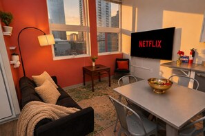 A modern living room with a sofa, a dining table, a TV mounted on the wall, and a view of the cityscape through large windows.