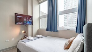 Habitació d'hotel amb llit, televisió penjada a la paret, finestra amb cortines i vista a la ciutat.