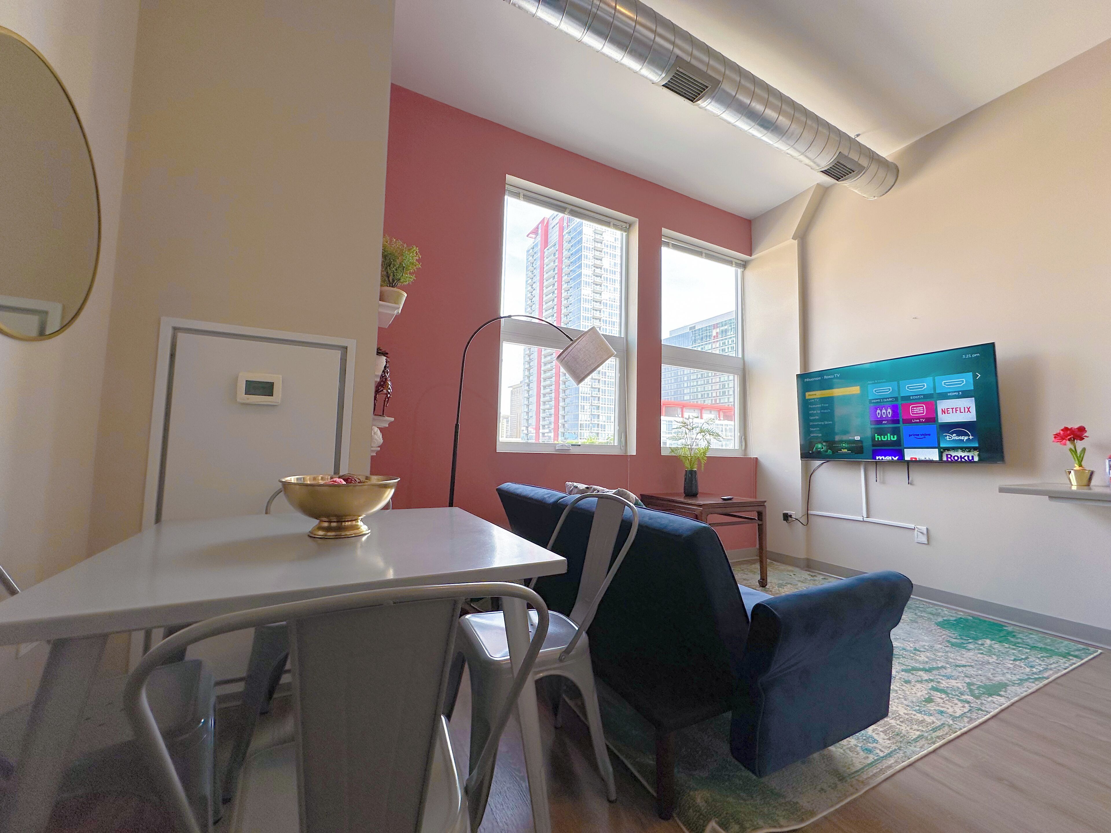 A modern living room with a dining area, a flat-screen TV mounted on the wall, and large windows offering a view of the cityscape.