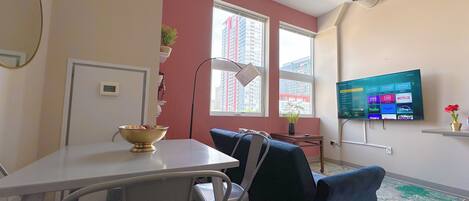 A modern living room with a dining area, a flat-screen TV mounted on the wall, and large windows offering a view of the cityscape.