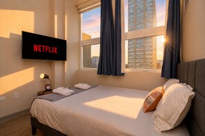 A hotel room with a bed, a TV mounted on the wall, a window with curtains, and a view of the cityscape.