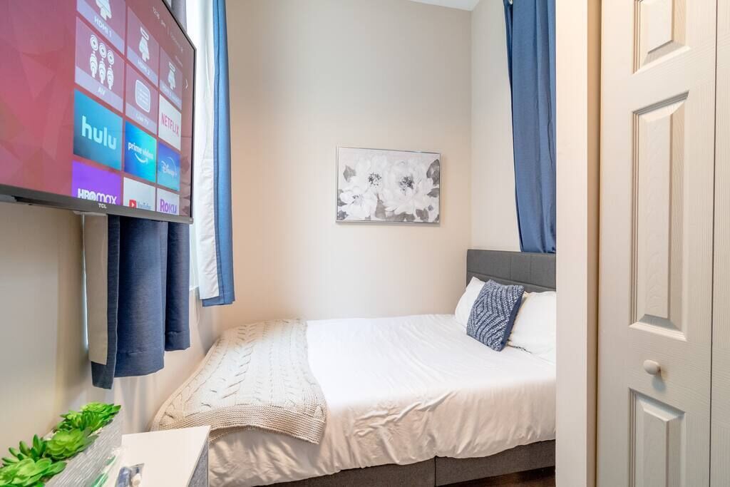 A neatly arranged bedroom with a bed, a wall-mounted TV displaying streaming services, a framed floral artwork, and a door.