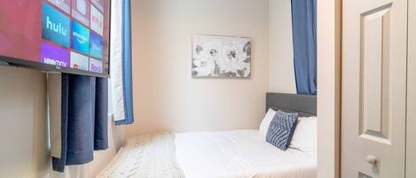 A neatly arranged bedroom with a bed, a wall-mounted TV displaying streaming services, a framed floral artwork, and a door.
