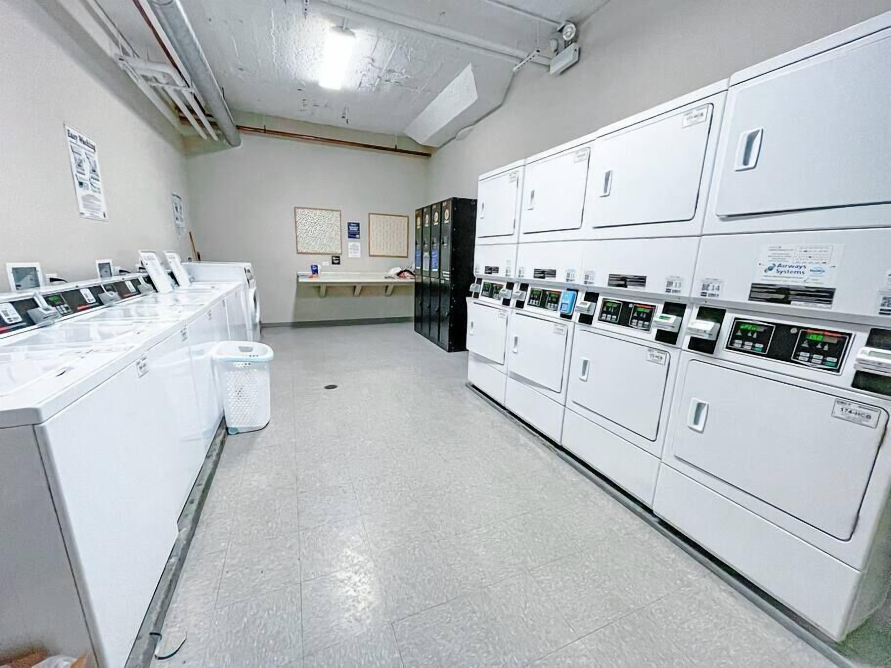 A laundry facility with multiple washing machines and dryers, a trash bin, and a bulletin board on the wall.