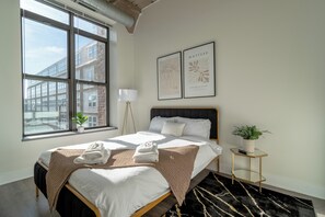 A modern hotel room with a large bed, a window with a city view, a bedside lamp, and two framed artworks on the wall. - Stylish 2BR Suite near McCormick Place & Soldier Field Best for Small Groups (Chicago)