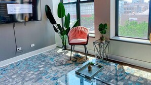 A modern office space with a large window, a pink chair, a glass coffee table, and a flat-screen TV on the wall.