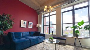 A modern living room with a blue sofa, a round glass coffee table, a potted plant, and a view of the city through large windows. - McCormick Place 2b/2b family unit with optional parking for up to 8 guests (Chicago)