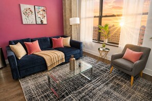 A living room with a dark blue sofa, a glass coffee table, a patterned rug, and a view of the city at sunset through large windows.