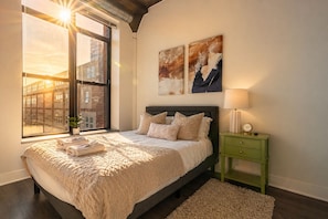 A bedroom with a large window, a bed with a beige blanket, a green nightstand with a lamp, and abstract wall art.