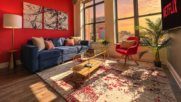 Un salon moderne avec un mur rouge, de grandes fenêtres, un canapé bleu, un fauteuil rouge et le logo de Netflix.