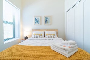 Ein ordentlich bezogenes Bett mit einer gelben Decke, weißen Kissen und einem gefalteten Handtuch seitwärts. Darüber an der Wand befinden sich zwei gerahmte Kunstwerke.