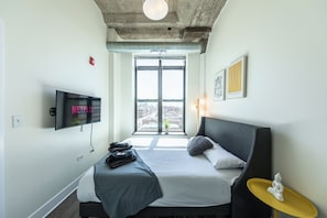 A modern hotel room with a large bed, a flat-screen TV, a view of the city, and a small table with a decorative item.