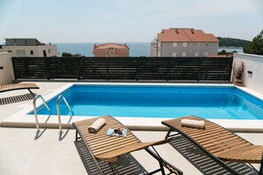 En pool på taket med solstolar i trä, en poolstege och utsikt över havet och byggnaderna.